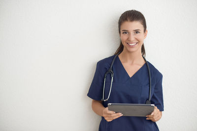 Portrait of smiling female doctor using digital tablet while standing against wall