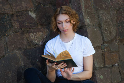 Young woman sitting on book against wall
