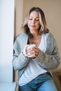 Young woman with blonde long curly hair in knitted grey sweater with cup of tea in hands at home