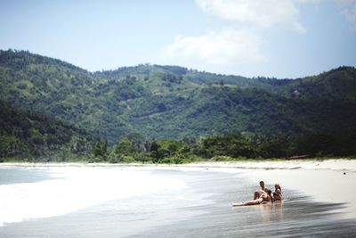 Couple sitting on mountain by sea against sky