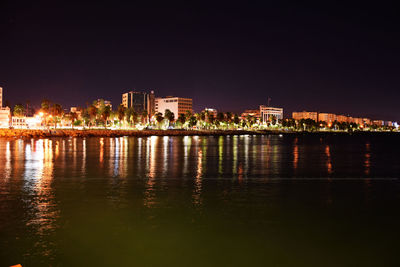 Calm river in front of illuminated buildings against sky at night