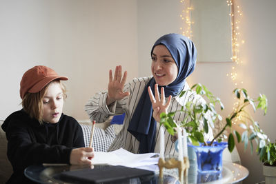 Mother wearing hijab holds up fingers while helping son doing math homework