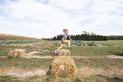 Young girl wearing a colorful dress jumping over hay bale at farm