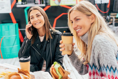 Two cheerful girlfriends are chatting and eating fast food at the street market.