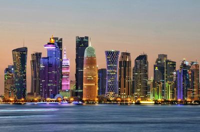 Illuminated buildings in city by sea against sky at dusk