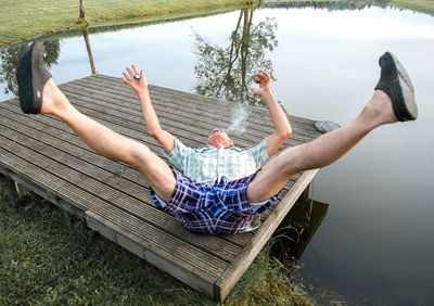 High angle view of man with feet up while smoking cigarette on pier at lakeshore