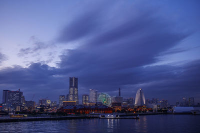 Illuminated buildings by river against sky at dusk