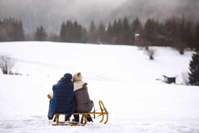 Back view of senior couple sitting side by side on sledge in snow-covered landscape