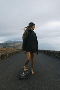 Carefree female hipster standing on longboard on asphalt roadway in highlands on lanzarote and looking away