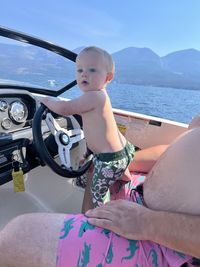 Baby boy standing on lap pretending to drive boat on a lake with mountains in background.