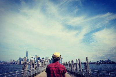 Rear view of man on pier leading towards city against cloudy sky