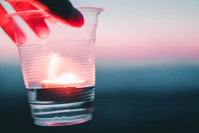 Cropped hand holding drink in disposable glass during sunset