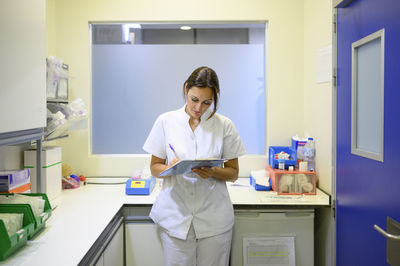 Woman in medical uniform writing on clipboard while standing in office during shift in children hospital