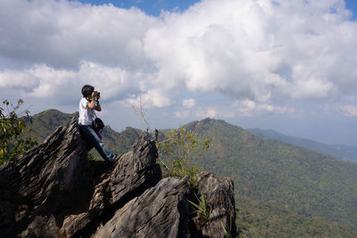 Woman sitting on rocks using camera to capture picture of mountains in summer