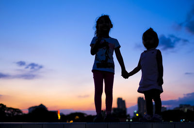 Low angle view of silhouette sisters standing on retaining wall against sky during sunset