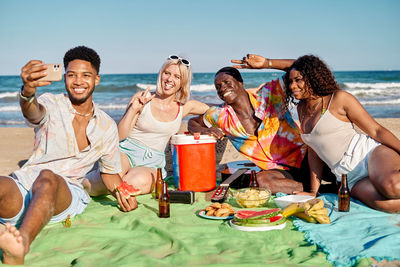 Group of cheerful young multiracial male and female friends in summer clothes smiling and showing two fingers gesture while taking selfie on smartphone during picnic on sandy beach