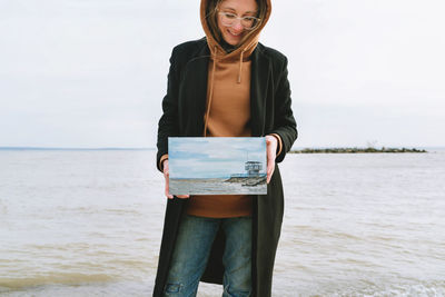 Smiling woman holding painting against sea