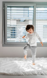 Full length of cute boy standing on bed by window at home