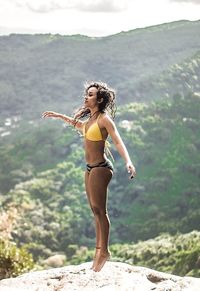 Side view of young woman in bikini levitating on cliff against mountains