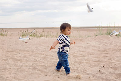 Baby boy in striped sailor t-shirt running on the sandy beach with seagulls near the sea in summer