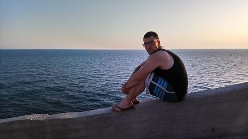 Young man sitting on sea against sky during sunset