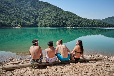 Back view of company of friendly people relaxing on tree log near lake and enjoying sunny day during vacation together