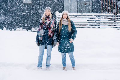 Two young women in warm winter clothes stand under the snowfall.
