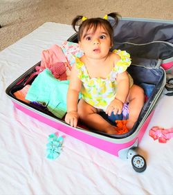 High angle view of cute baby girl sitting in suitcase on floor at home