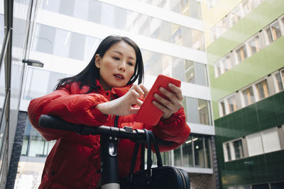 Female commuter using smart phone while leaning on electric push scooter handlebar in city