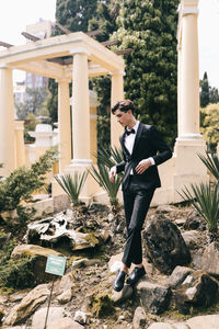 A beautiful young man, the groom in an elegant wedding suit, stands posing in the city's old park