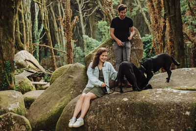 Young couple with two dogs having fun in forest while sitting on rock