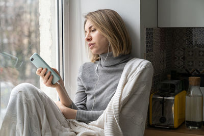 Bored female addicted and chatting in social media, sitting on windowsill at home using mobile phone