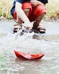 Low section of boy with paper boat splashing water at lakeshore