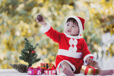 Full length of cute baby girl wearing santa costume while sitting with christmas decorations on table against trees