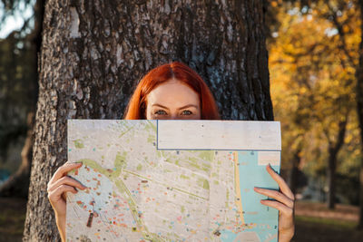 Mid adult woman holding map in front of face against tree in park