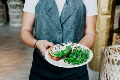 From above crop cook holding plate with tasty big appetizing sandwich with avocado sauce and greens