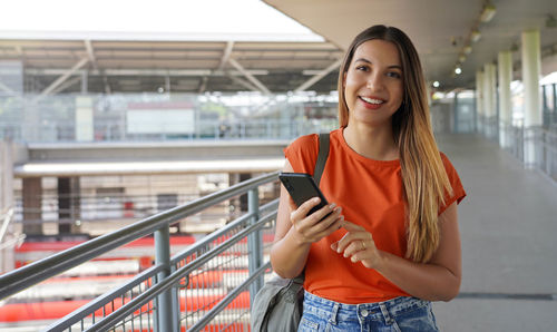 Positive trendy female student using mobile phone at train station. 