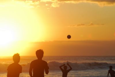 Friends playing beach volleyball against orange sky during sunset