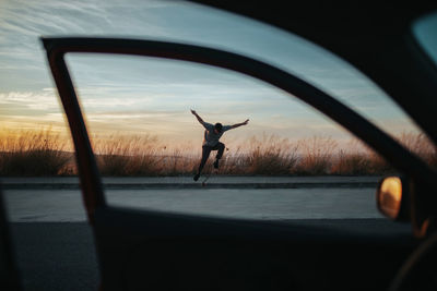 From car view of full body young man in casual wear jumping on skateboard while performing kickflip on asphalt road against dusky sky