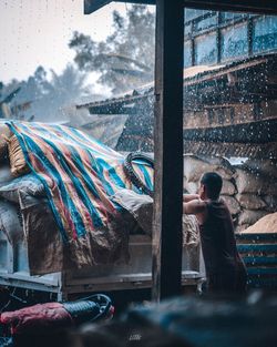Rear view of man standing by window in rainy season