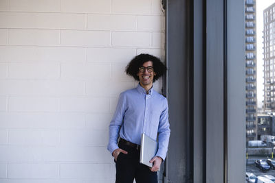 Happy businessman holding laptop standing with hand in pocket in office