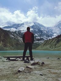 Rear view of man standing at lakeshore against snowcapped mountains