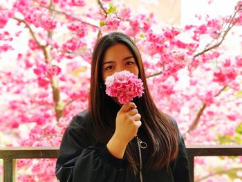 Portrait of young woman holding pink flowers against tree