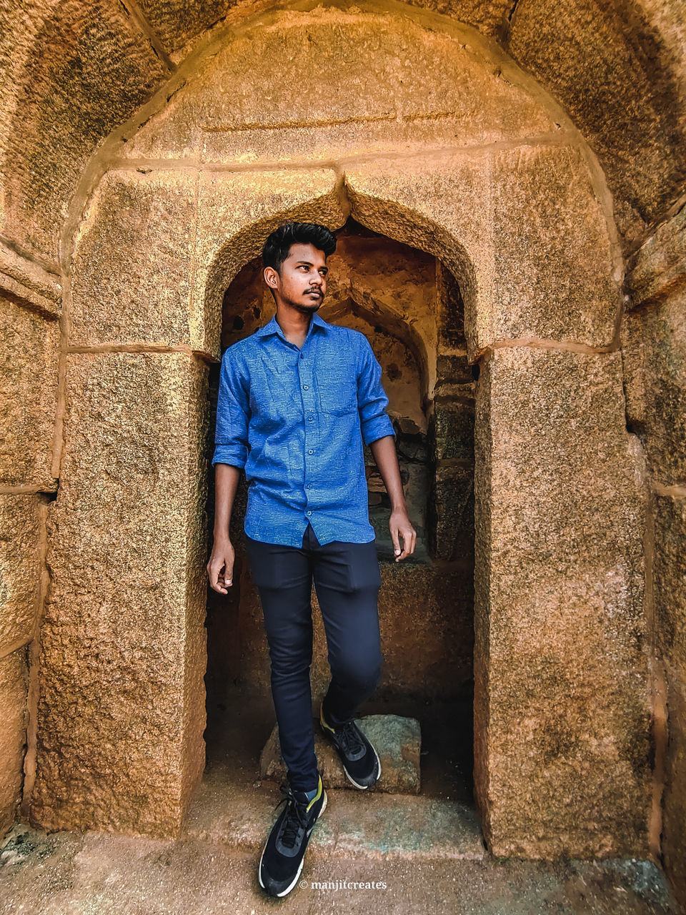full length, one person, architecture, casual clothing, front view, adult, standing, portrait, temple, arch, ancient history, young adult, men, built structure, day, hands in pockets, looking at camera, jeans, leisure activity, lifestyles, smiling, history, blue, clothing, glasses, the past, footwear, fashion, looking, outdoors, building exterior, cave, rock, person