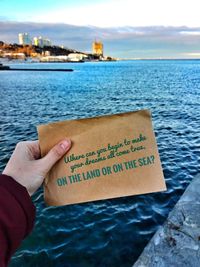 Hand holding text by sea against sky