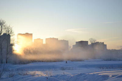 Buildings on snow covered landscape against sky during sunset