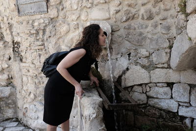 Side view of woman drinking water from fountain against stone wall