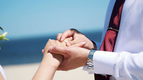  the bride and groom wear rings to each other, against the background of the sea, sand and blue sky.