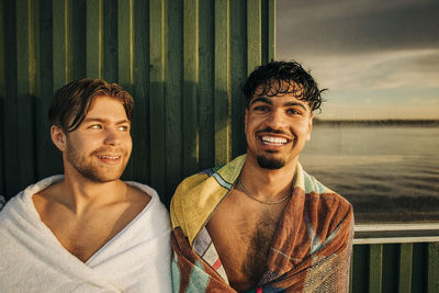 Portrait of happy man wearing towel while sitting with male friend against cottage