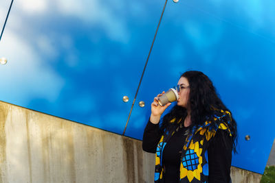 Woman with long curly hair drinks coffee from paper cup on background of blue wall